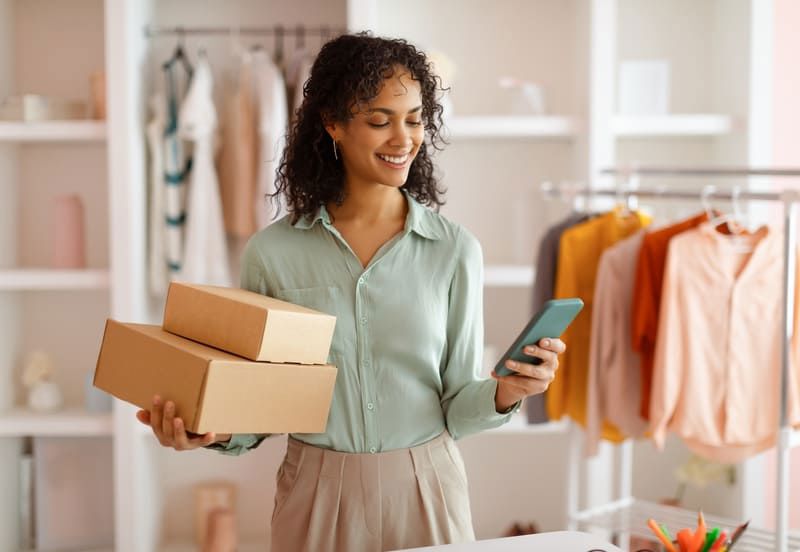 Retail and e-commerce businesswoman smiling confidently while holding boxes and her phone.