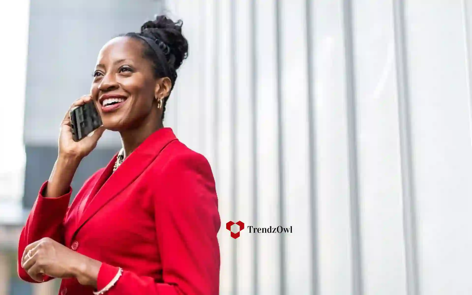 A person in a red suit speaks on a phone, smiling. they are standing outdoors beside a striped white background. text on the image reads "trendzowl."