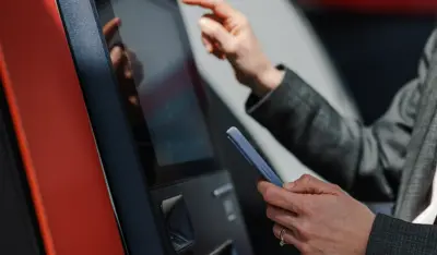 Person using a self-service banking kiosk while holding a smartphone, highlighting TP's digital banking solutions.