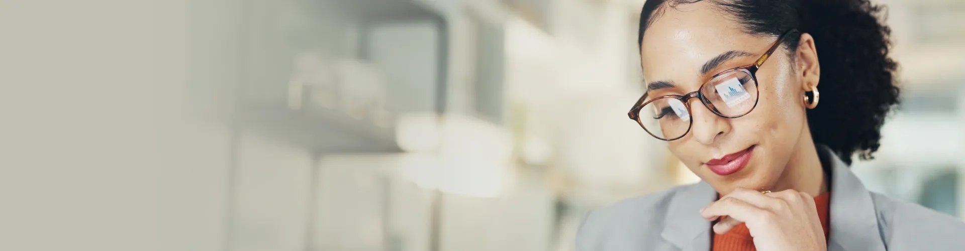 A professional woman thoughtfully analyzes data, with charts reflected in her glasses in a bright office.