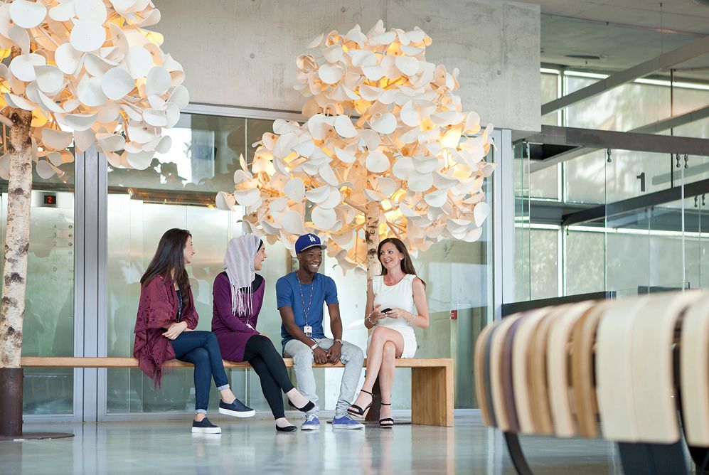 A group of four colleagues chatting while seated on a wooden bench in a modern office lobby with decorative white hanging lights and large glass windows.